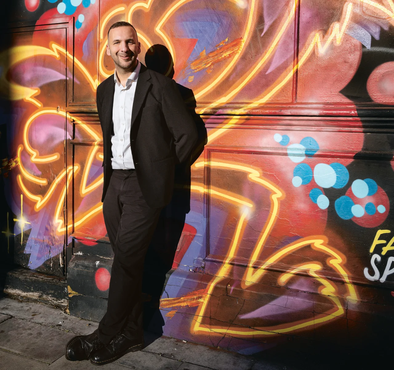 Zack Polanski in schwarzem Anzug lehnt lächelnd vor bunter Wand mit leuchtendem Graffiti