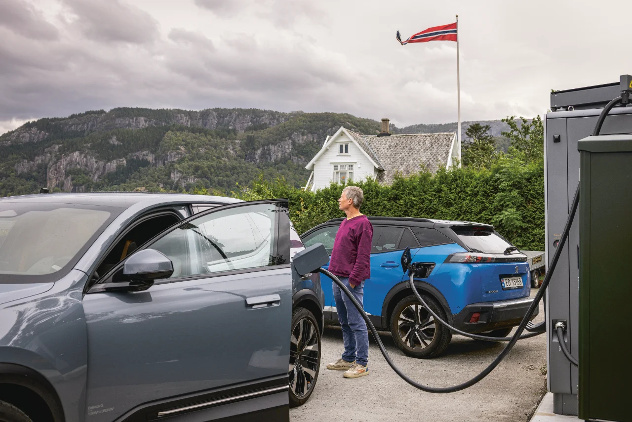 Mann lädt sein Elektroauto an einer Ladesäule in Norwegen, im Hintergrund zu sehen ist ein weißes Holzhaus mit norwegischer Flagge und ein Bergpanorama