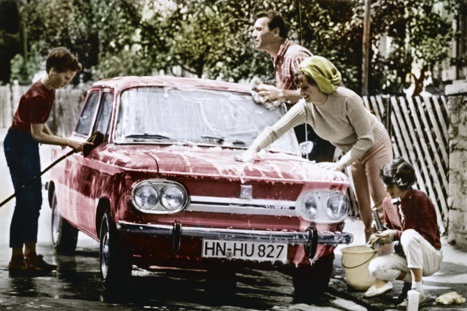 Ein historisches Farbfoto aus den 1960er Jahren zeigt eine vierköpfige Familie, die gemeinsam ein rotes NSU Prinz Oldtimer-Fahrzeug auf einer Einfahrt wäscht.