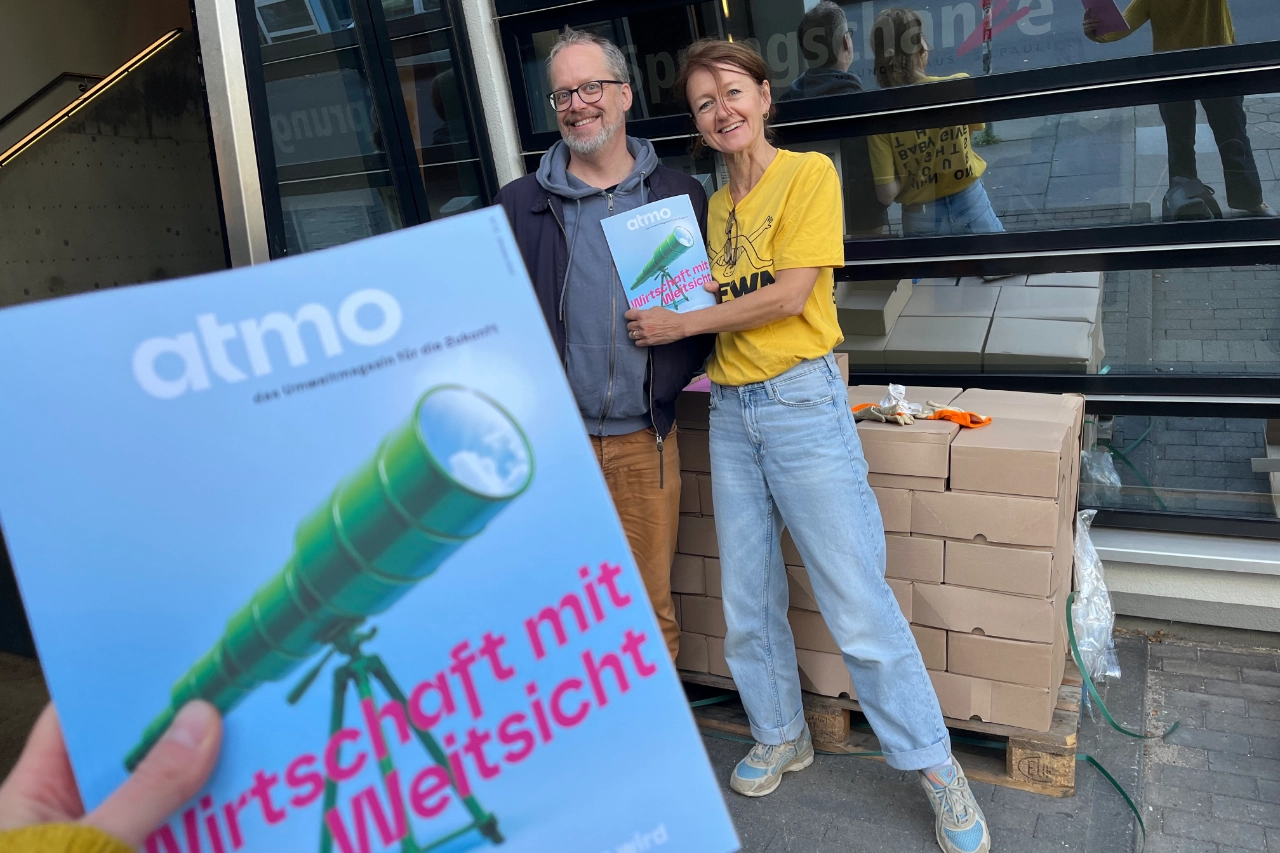 Im Vordergrund hält eine Hand das Magazin „atmo“ mit dem Titel „Wirtschaft mit Weitsicht“. Im Hintergrund lächeln zwei Personen vor gestapelten Kartons.