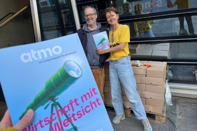 Im Vordergrund hält eine Hand das Magazin „atmo“ mit dem Titel „Wirtschaft mit Weitsicht“. Im Hintergrund lächeln zwei Personen vor gestapelten Kartons.