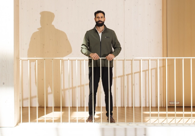 Ein Mann mit Bart steht hinter einem weißen Geländer in einem modernen Holzgebäude.