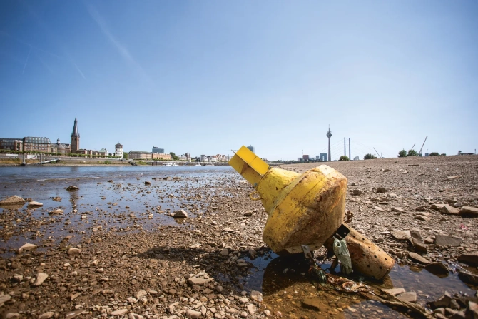 Eine gelbe Boje liegt auf dem trockenen, steinigen Rheinufer bei extremem Niedrigwasser, im Hintergrund eine Stadtkulisse