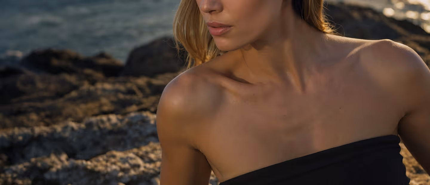 Close-up of a woman wearing a black strapless top, with ocean and rocky background.