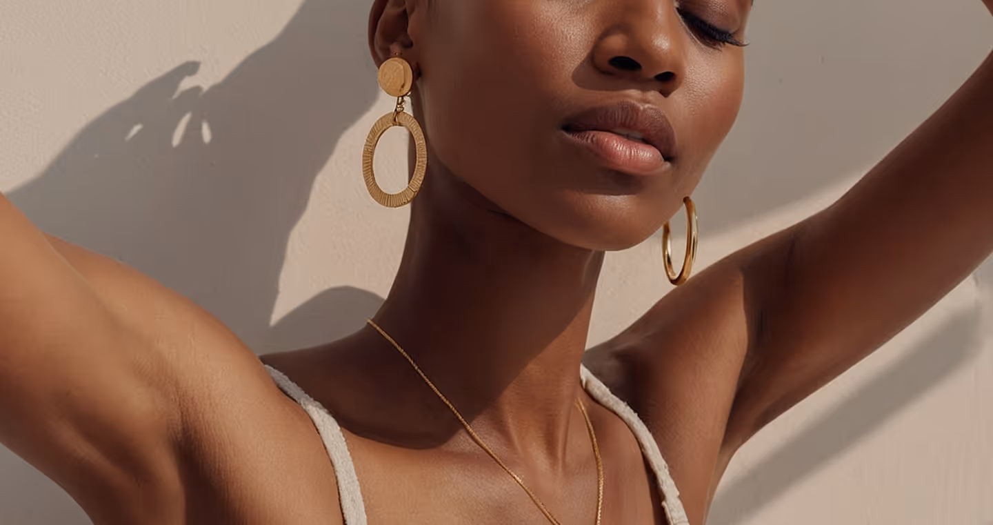 Close-up of a woman with eyes closed wearing gold hoop earrings and a pendant necklace against a beige background.