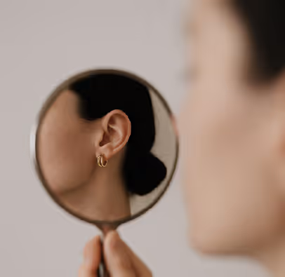 Close-up of a woman's ear reflected in a handheld round mirror, showing a gold hoop earring.