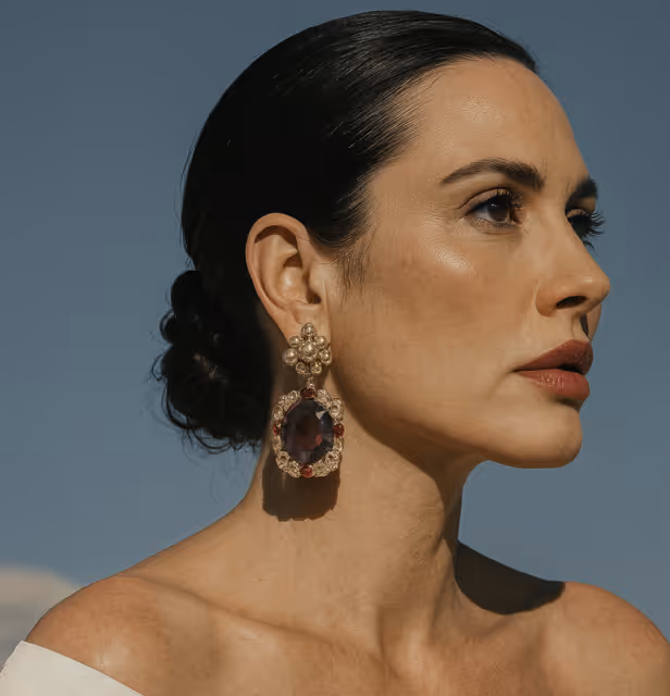 Profile of a woman with dark hair in a low bun, wearing large ornate earrings and a white off-shoulder top against a clear sky background.