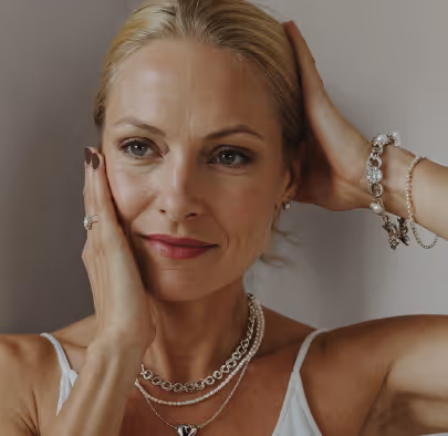 Blonde woman with styled hair wearing layered necklaces, bracelets, and a ring, gently touching her face and head.