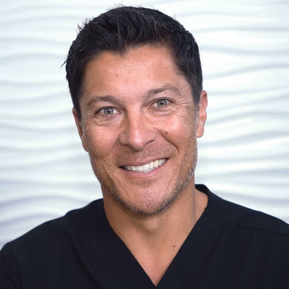 Smiling man with short dark hair wearing a black shirt against a white wavy patterned background.