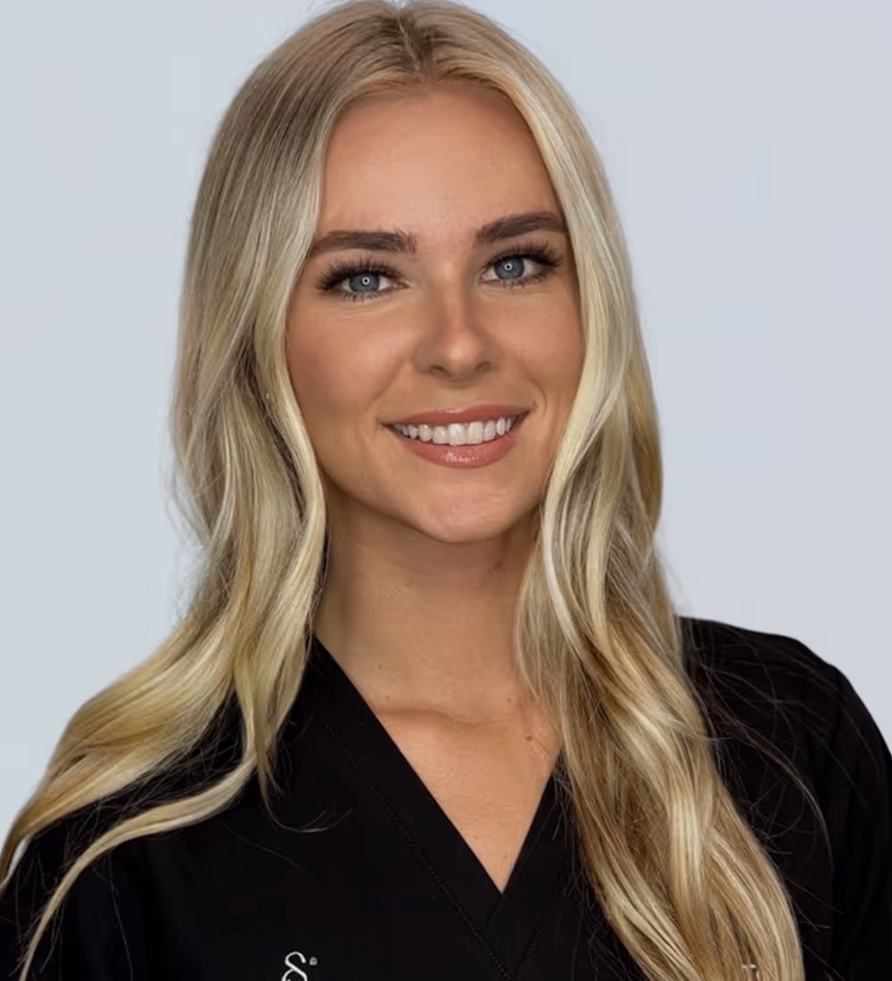 Smiling young woman with long blonde hair wearing a black V-neck top against a plain light background.