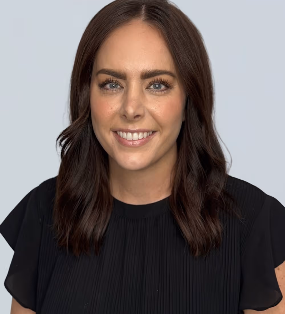 Smiling woman with shoulder-length dark brown hair and blue eyes wearing a black top against a light gray background.