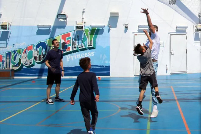 Children and adults play basketball on the sports deck of the Discovery Princess, framed by the colorful postcard mural featuring world landmarks and bold Discovery lettering.

