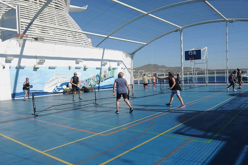Guests play pickleball on the sports court of the Discovery Princess cruise ship, with the vibrant “Greetings from Discovery Princess” mural in the background creating a lively and shareable backdrop.
