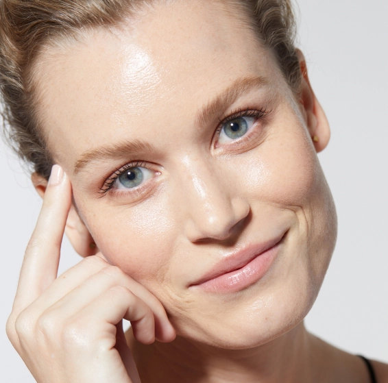 A woman with light hair and blue eyes smiles gently, touching her face with one hand.