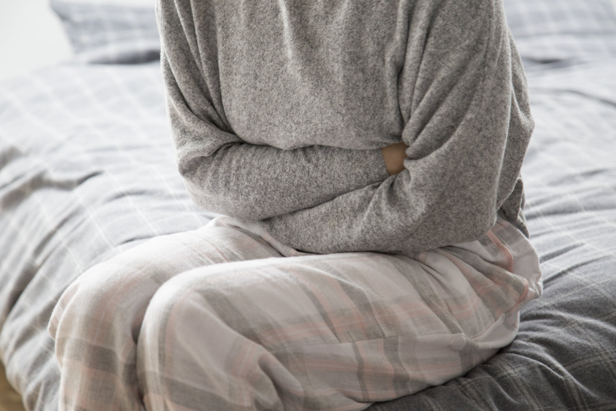 A person in a grey sweater and plaid pants sits on a bed, arms crossed over their abdomen, suggesting chronic discomfort.