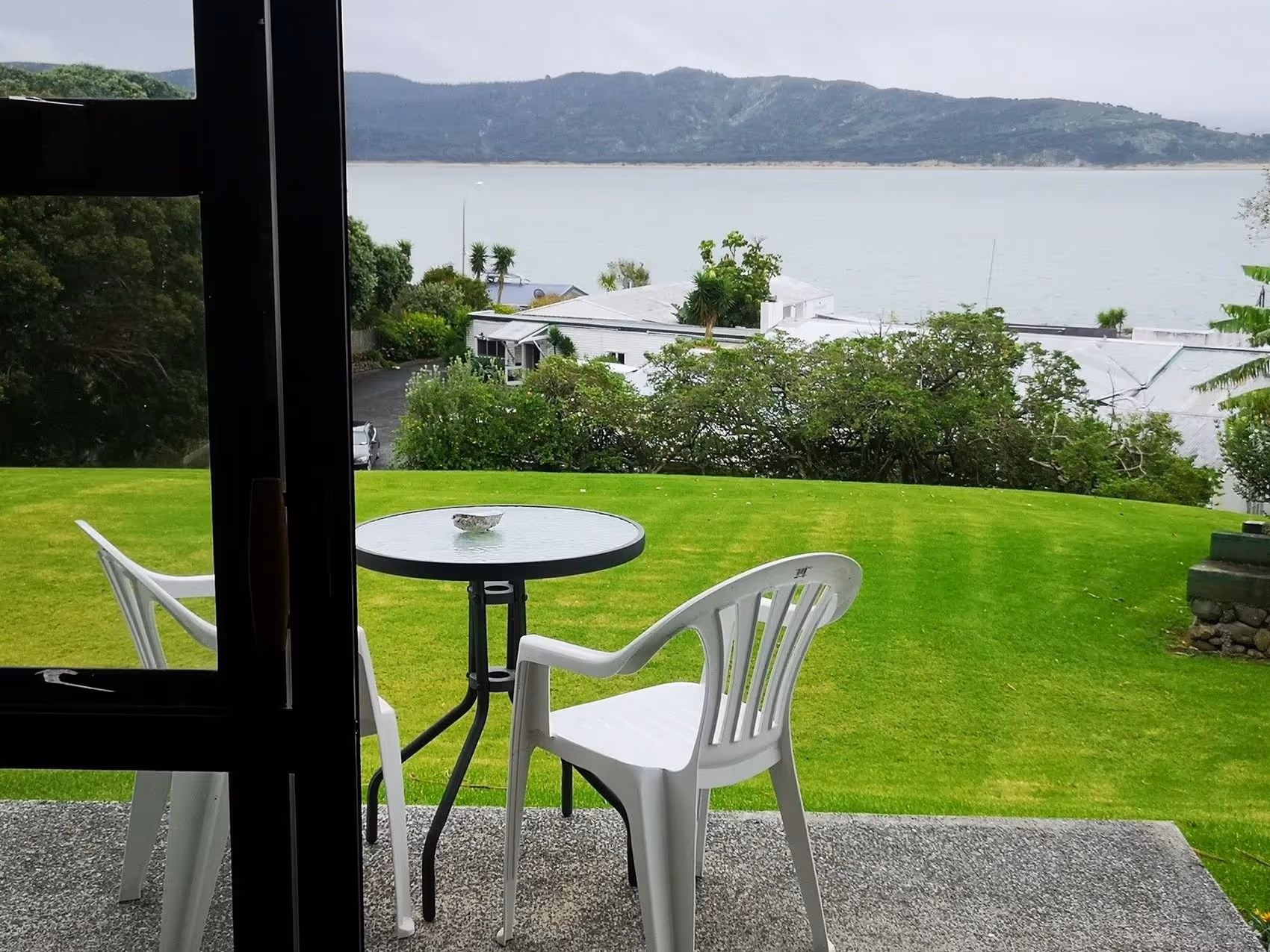 A cozy outdoor seating area with a round table and two white chairs, overlooking a serene lake and rolling green hills in the background.