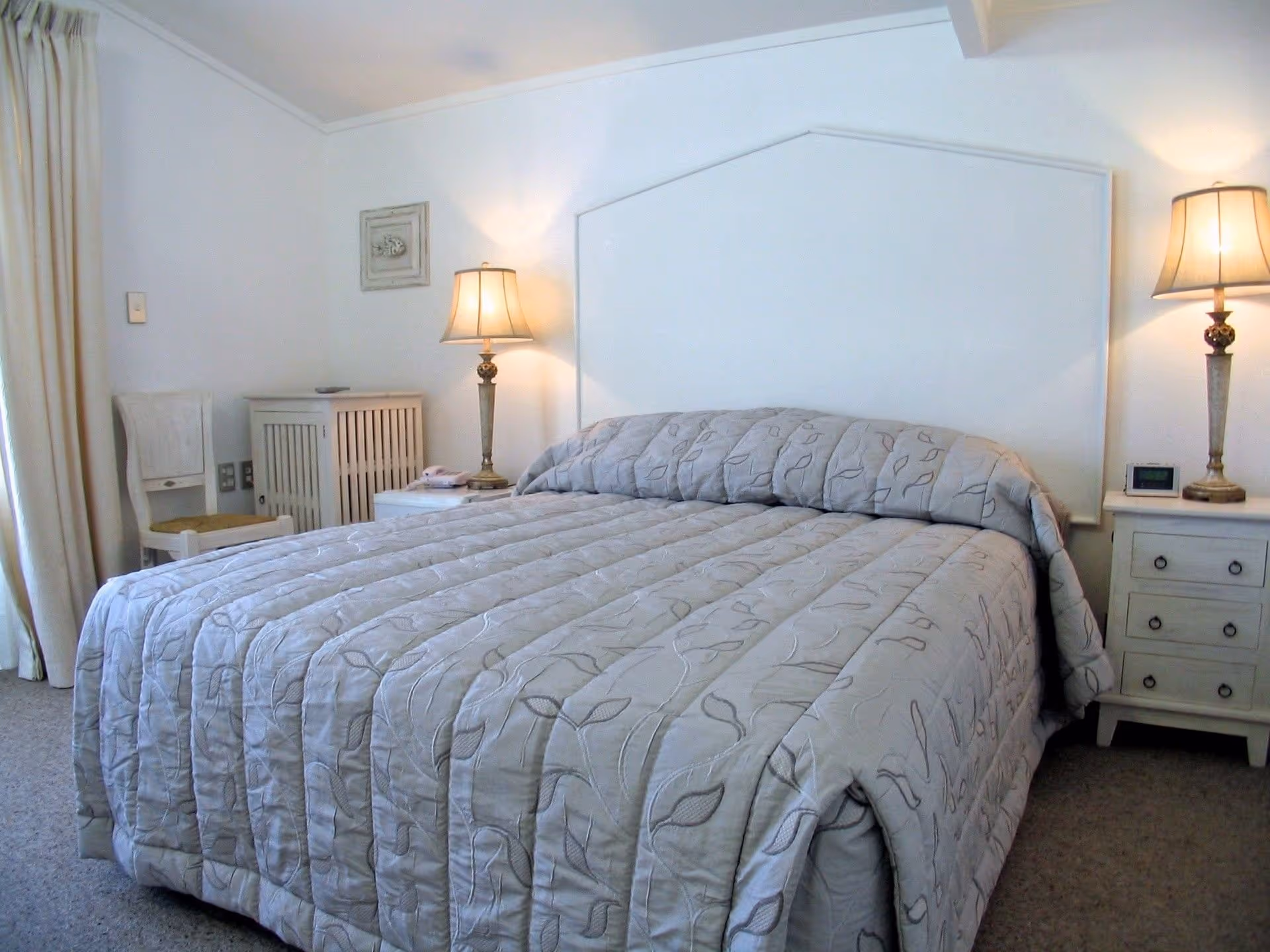 Cozy bedroom featuring a queen bed with a gray quilt, nightstands, lamps, and cream curtains, creating a serene atmosphere.
