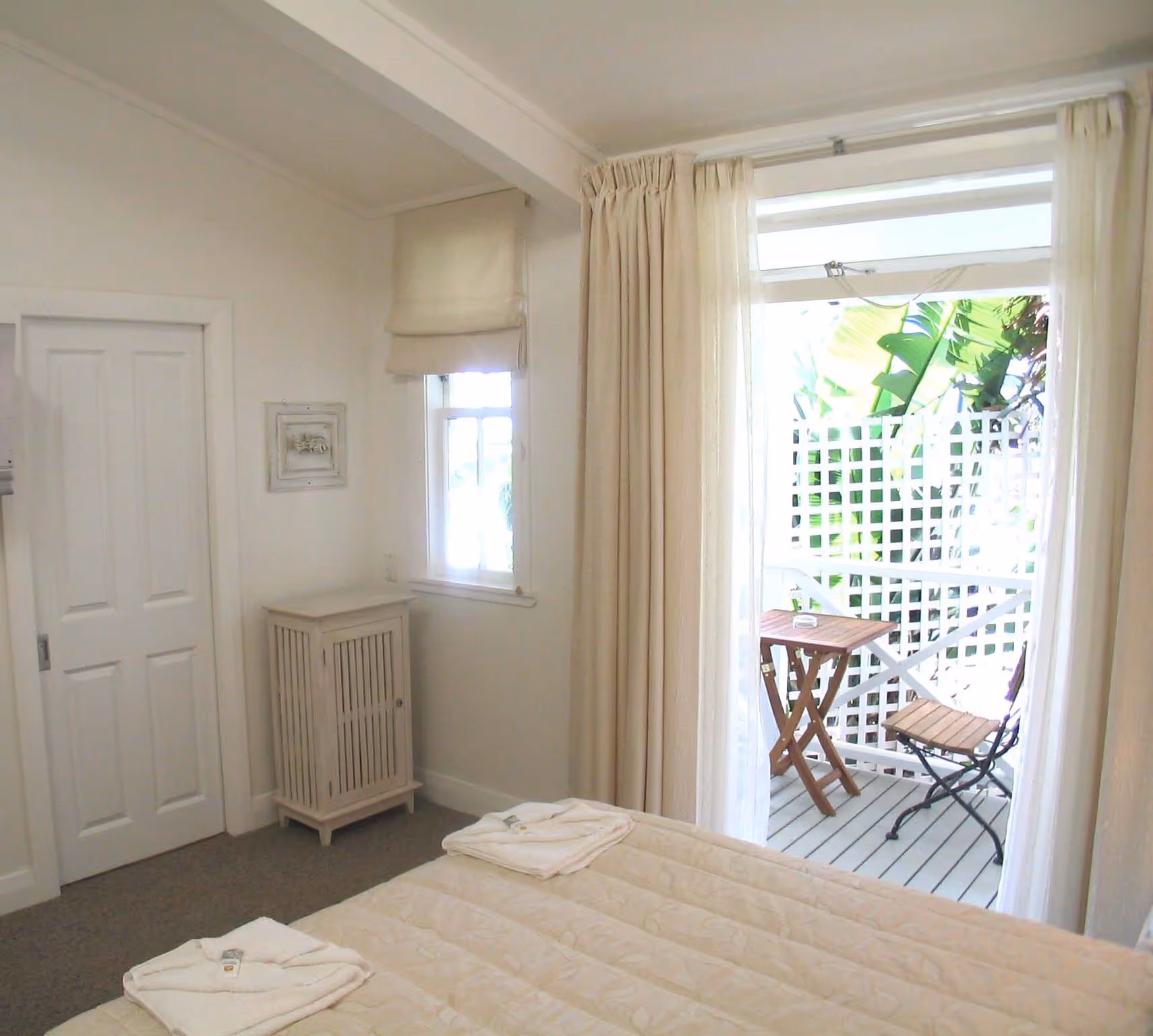 Bright bedroom with a white bedspread, plush towels, and curtains revealing a small patio with a table and chairs surrounded by greenery.