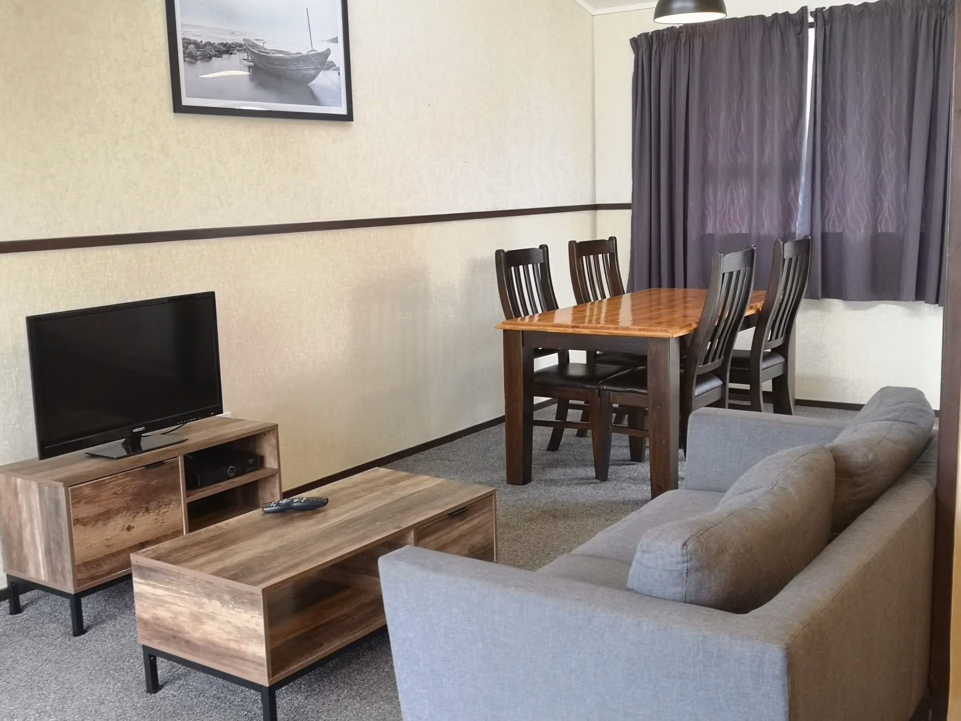 Cozy living area featuring a gray sofa, wooden dining table with chairs, TV on a rustic media stand, and sheer curtains.