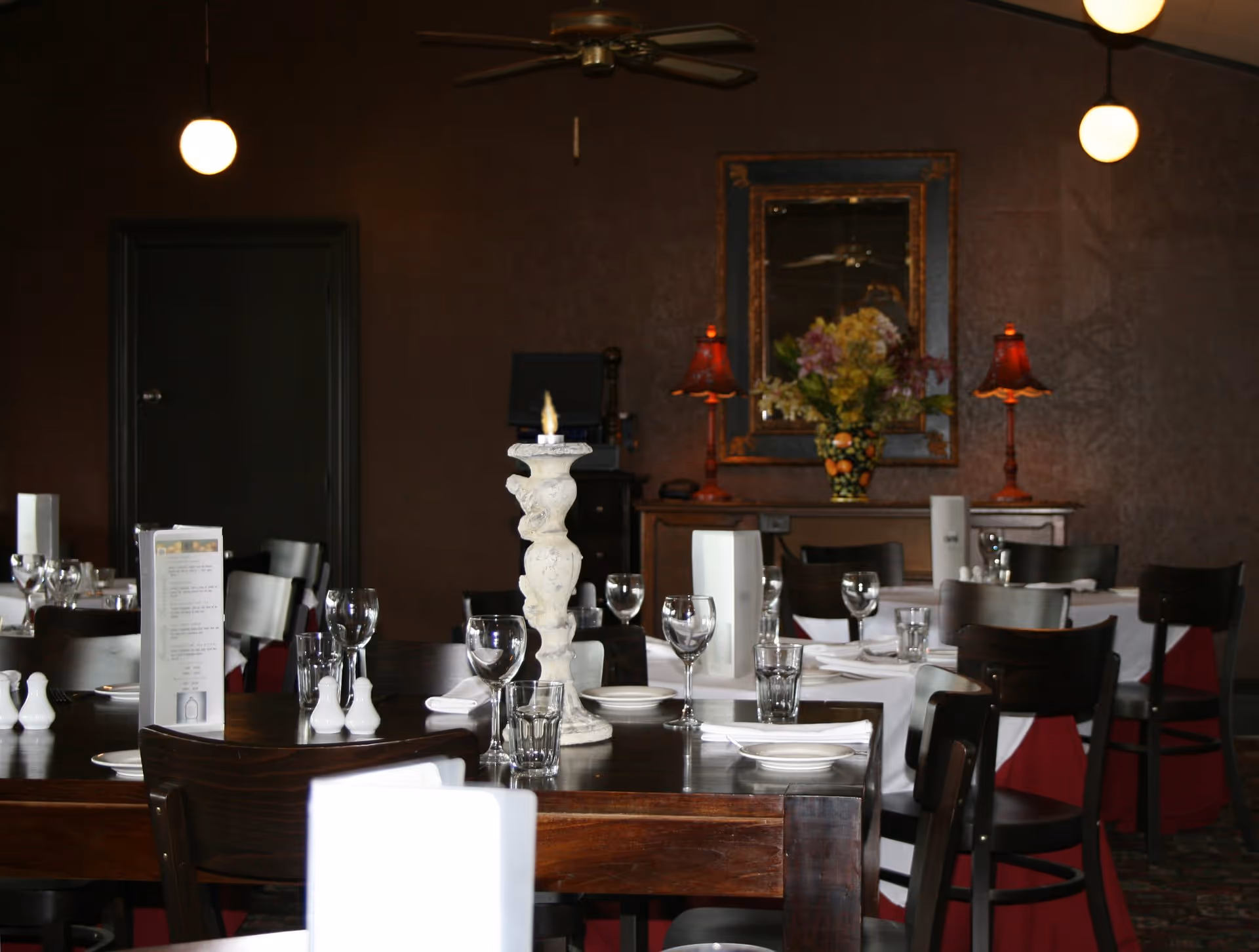 Elegant restaurant interior featuring dark walls, wooden tables, white tablecloths, and decorative lamps with floral arrangements.