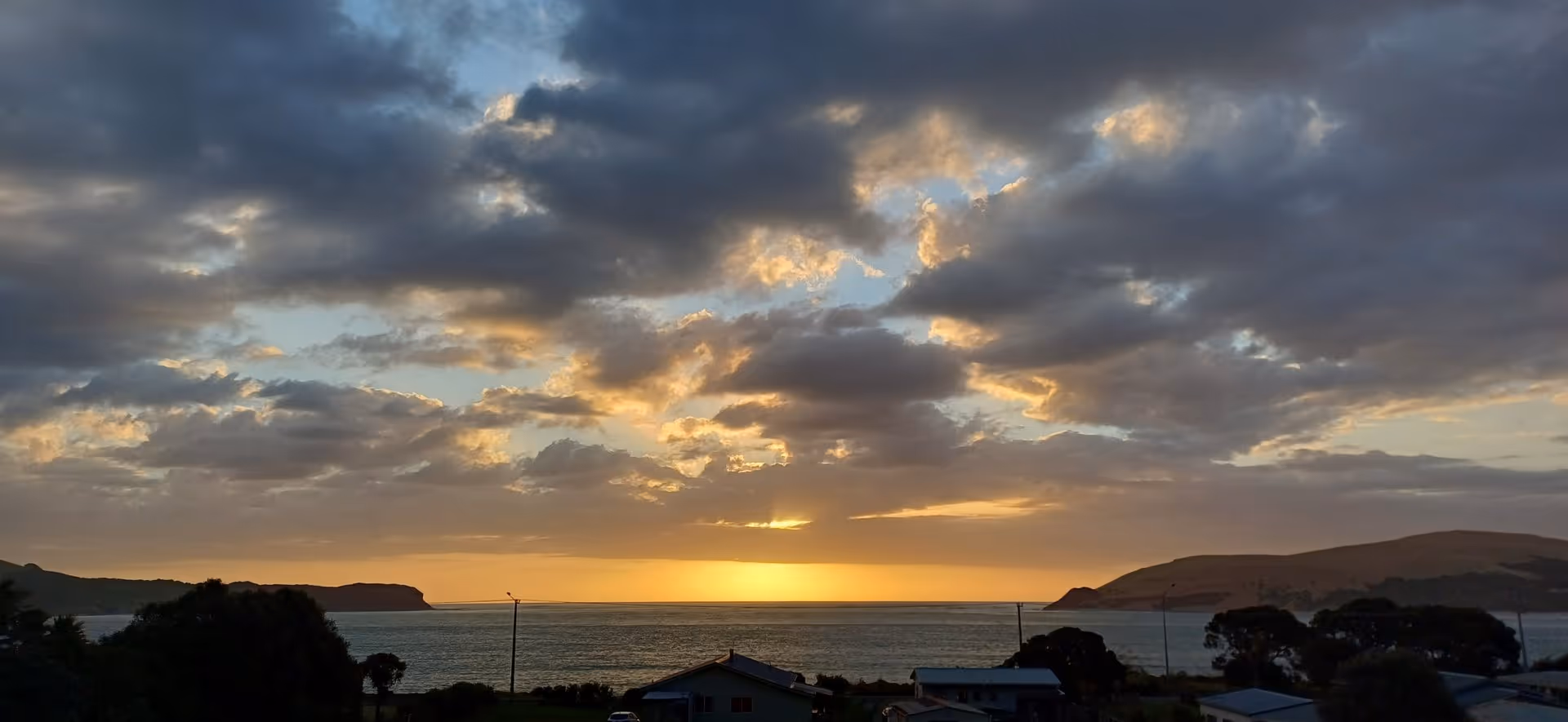A serene sunset over a calm ocean, framed by dark clouds and distant hills, with houses silhouetted along the shoreline.
