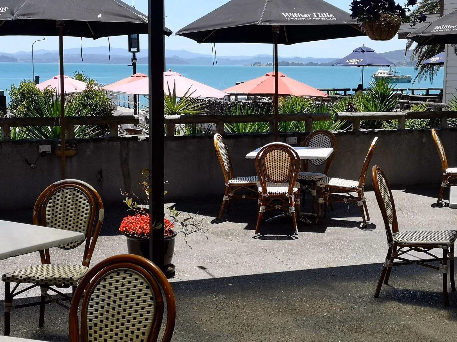 Outdoor café with wicker chairs and tables, surrounded by tropical plants, umbrellas, and a scenic view of the blue water and mountains.