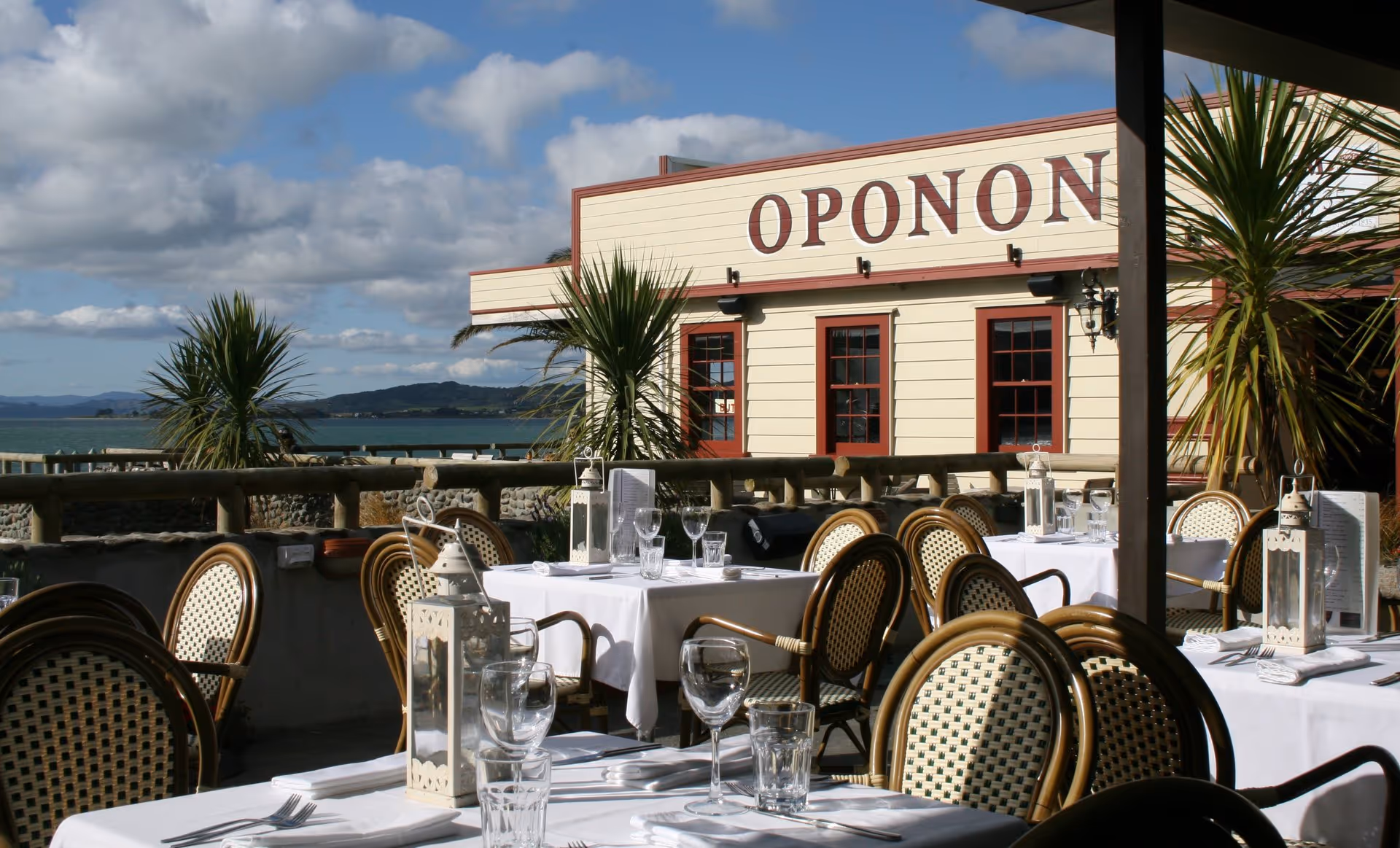 Outdoor dining area of the Opononi restaurant, featuring elegant tables set with glassware, overlooking the ocean and lush greenery.