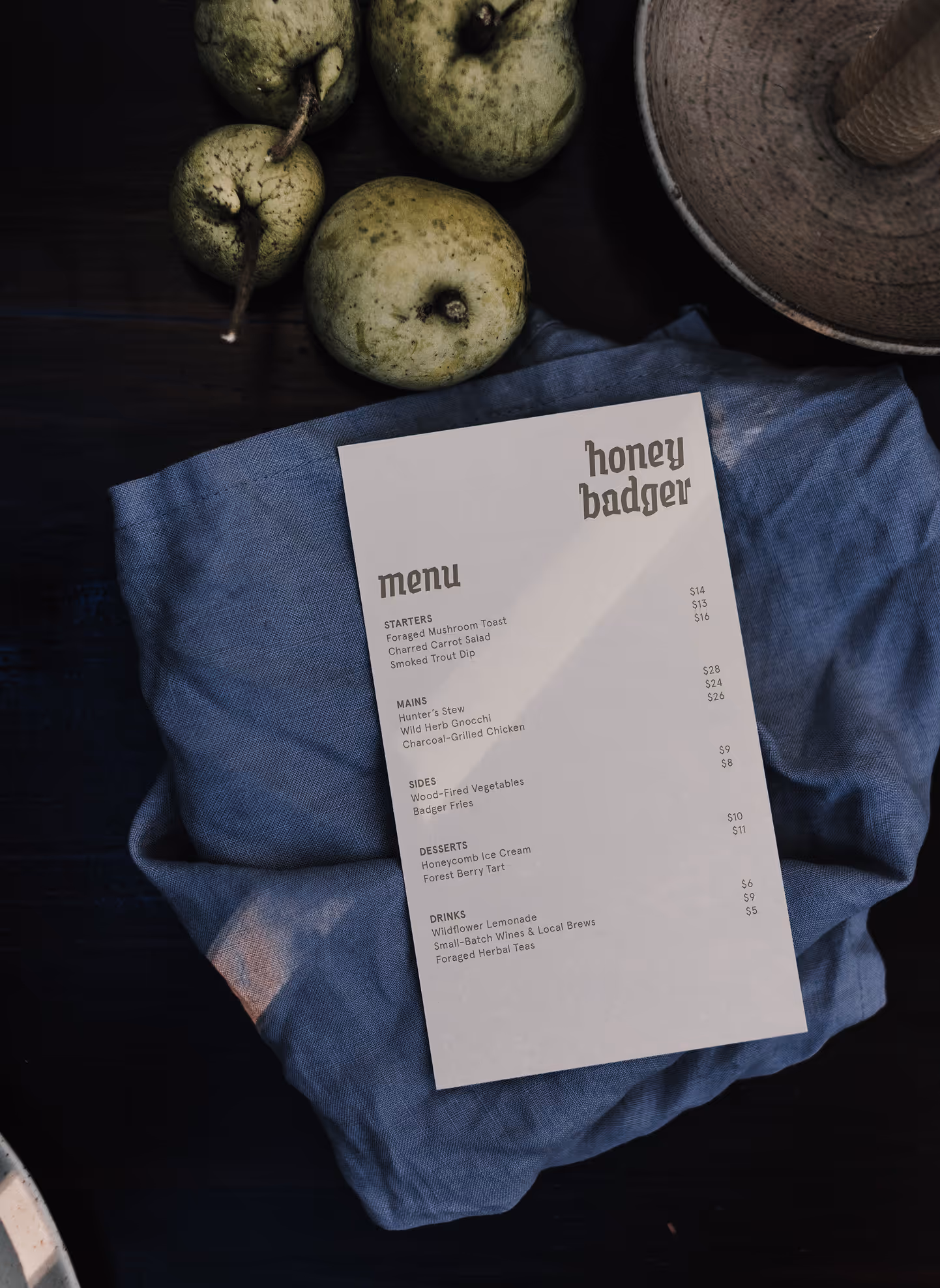 A printed Honey Badger restaurant menu placed on a blue cloth, surrounded by fresh green pears and natural tableware.