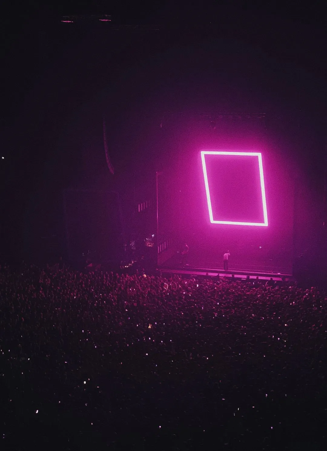 Wide shot of a concert stage with a bright pink square light and a large crowd in silhouette, captured in a dark venue.