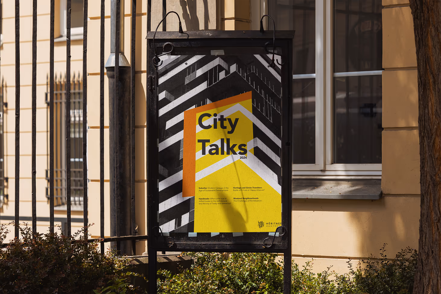 City Talks poster displayed in a black metal frame on a city sidewalk, featuring bold geometric patterns and yellow, orange, and black design elements.