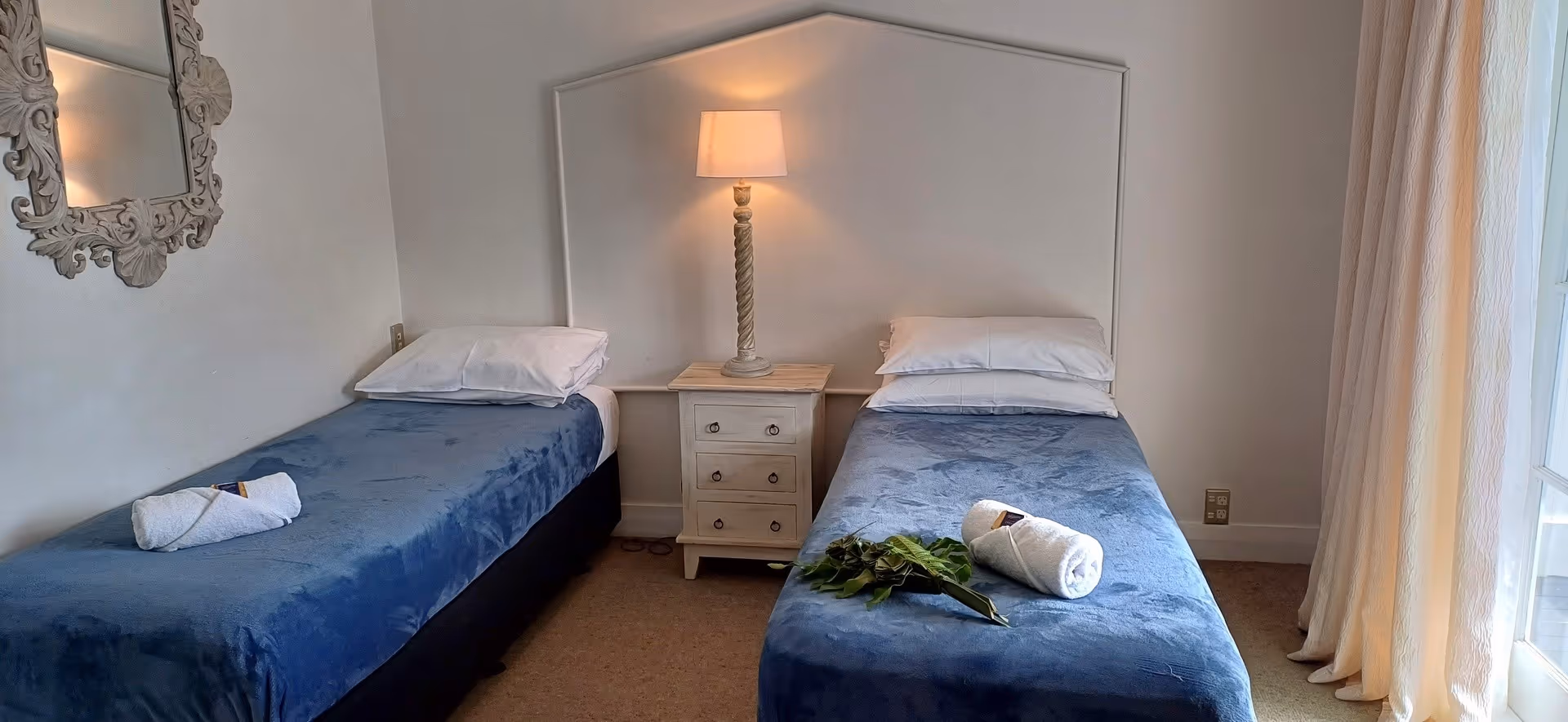 Bright, airy bedroom with two twin beds, a nightstand, and a modern lamp, featuring soft blue bedding and minimal decor.