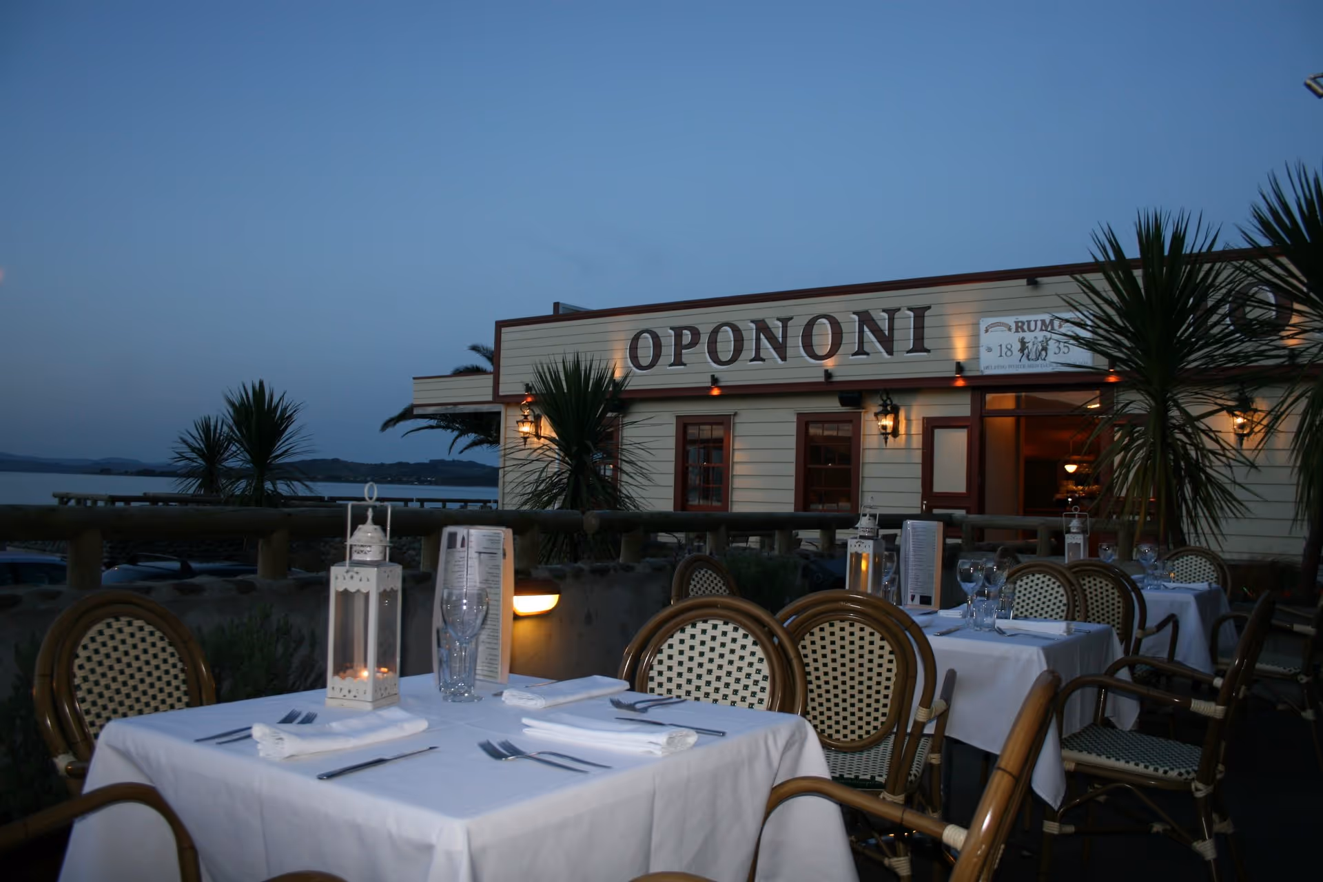 A seaside restaurant at dusk, featuring elegantly set tables with chairs, palm trees, and a charming facade that reads "Onononi".