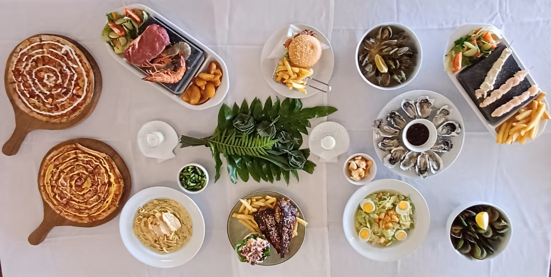 A stylishly arranged spread of diverse dishes, including pizza, pastries, salads, and dips, centered around fresh green garnishes.