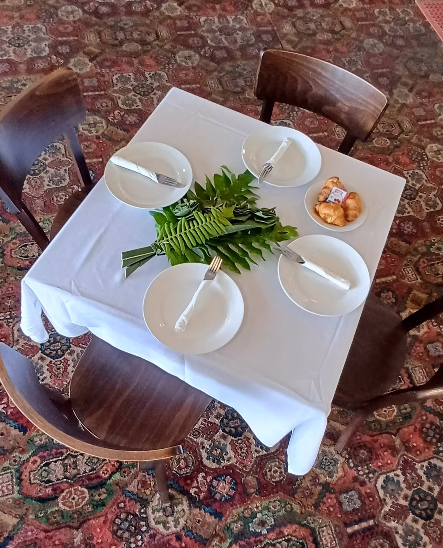 A neatly set dining table with four plates, cutlery, and a centerpiece of green vegetables, surrounded by wooden chairs.