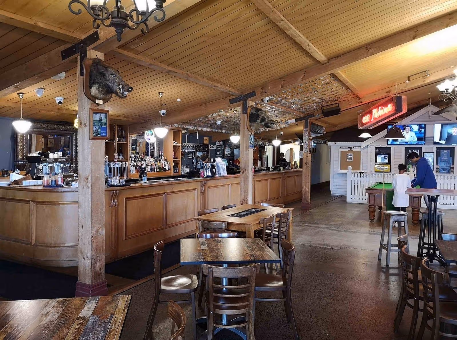 A cozy wooden bar interior featuring high ceilings, rustic decor, and empty tables, with patrons engaged at the bar in the background.
