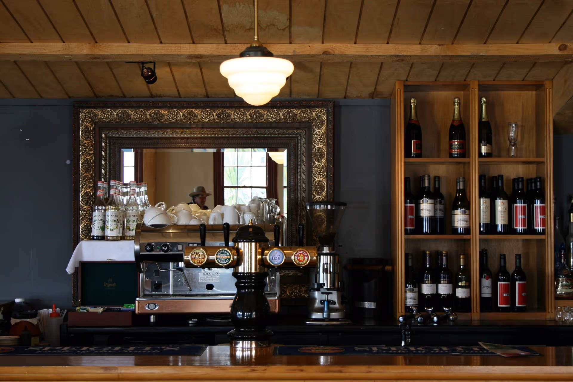 Cozy bar interior featuring a wooden counter, vintage mirror, shelving with various bottles, and warm overhead lighting.