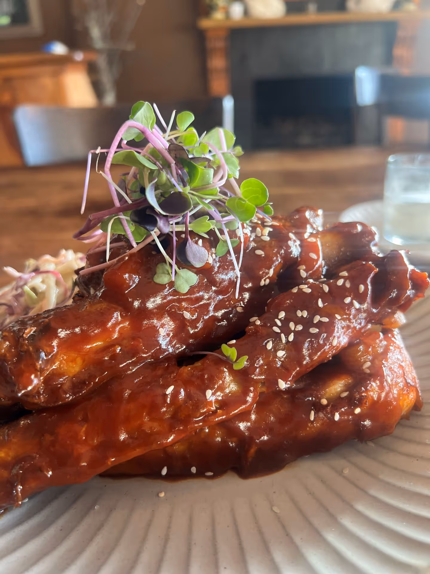 A plate of sticky, glazed chicken wings topped with fresh microgreens and sesame seeds, set against a blurred background.
