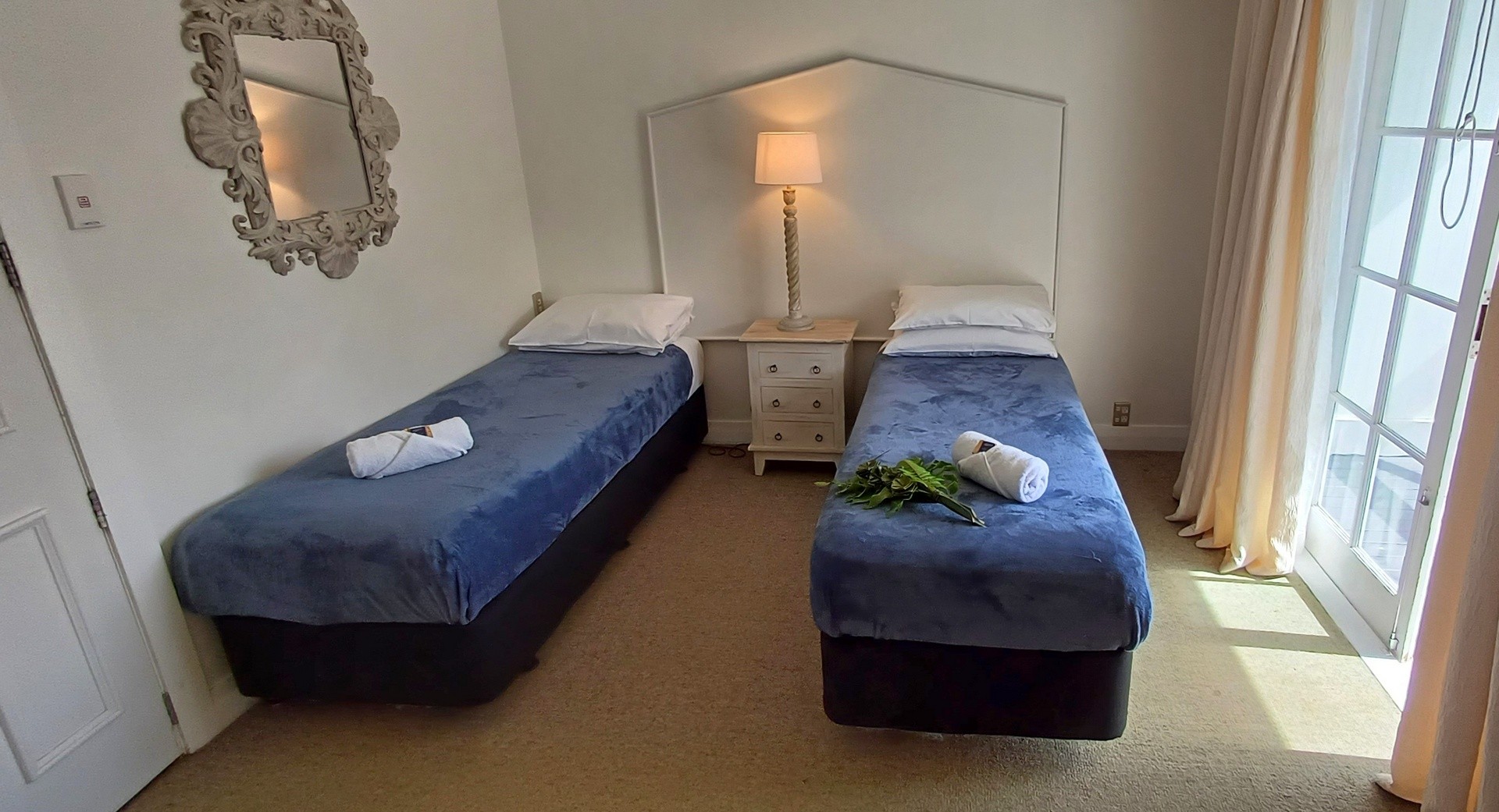 Cozy guest room featuring two twin beds with blue bedding, a decorative mirror, a floor lamp, and natural light from a window.