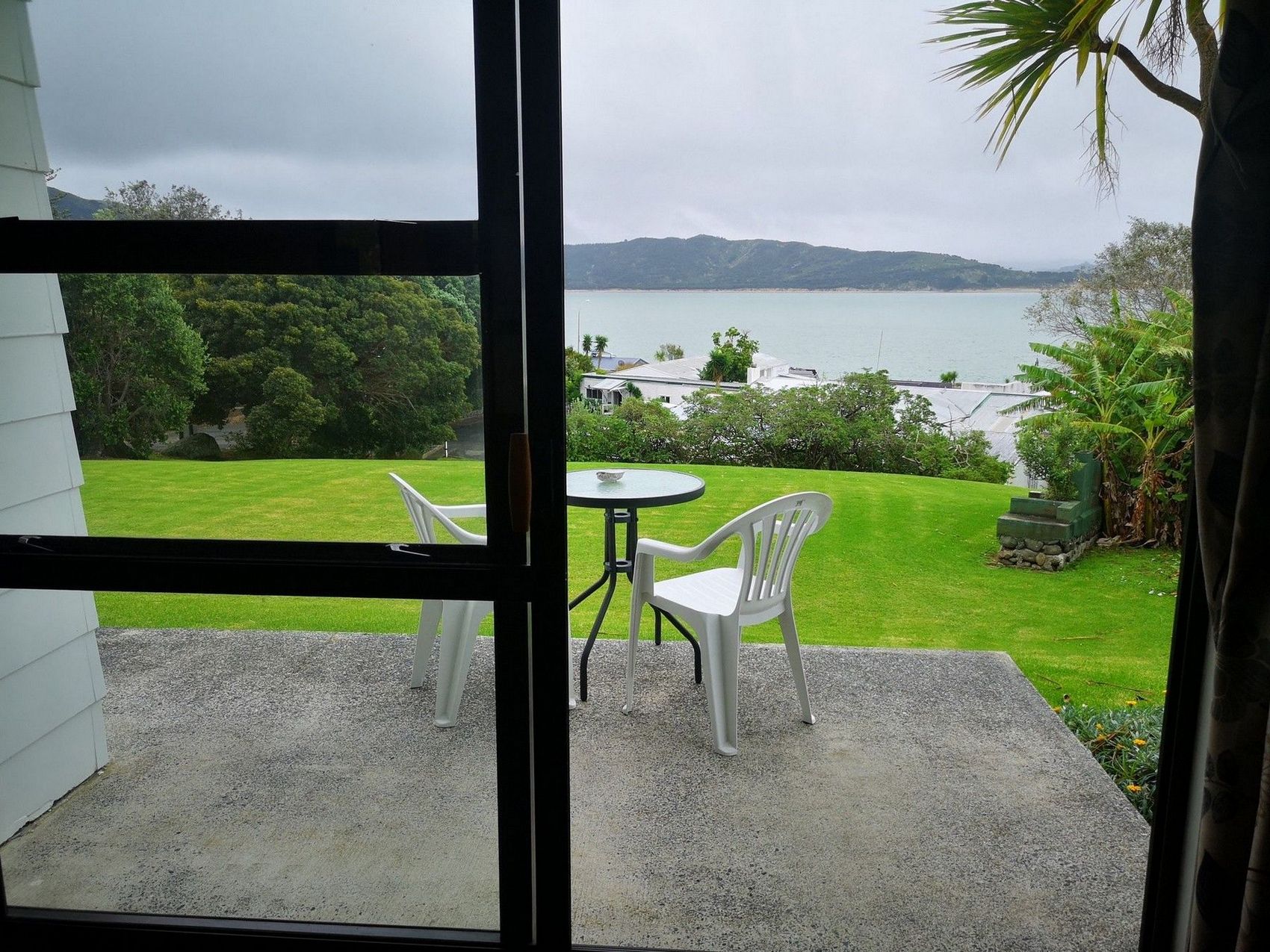View through a glass door of a patio with a table and chairs, overlooking a grassy area and a scenic bay under cloudy skies.