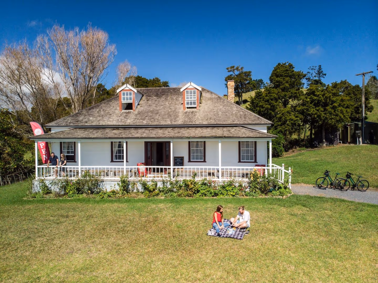 Aerial view of a charming, historic house with a porch, surrounded by greenery, where two people enjoy a picnic on the lawn.