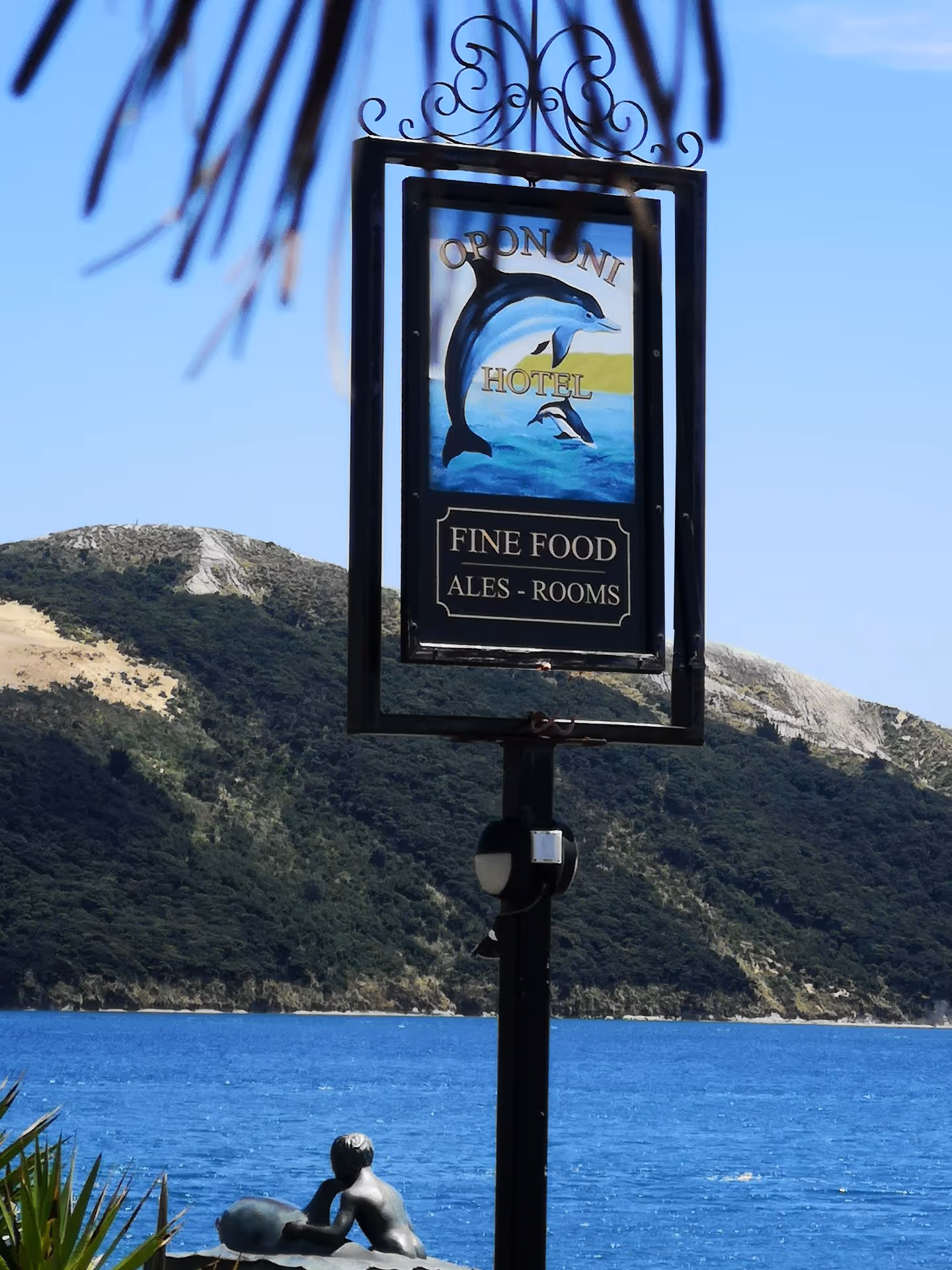 A picturesque sign for Opononi Hotel stands by a tranquil blue lake, framed by lush greenery and distant hills.