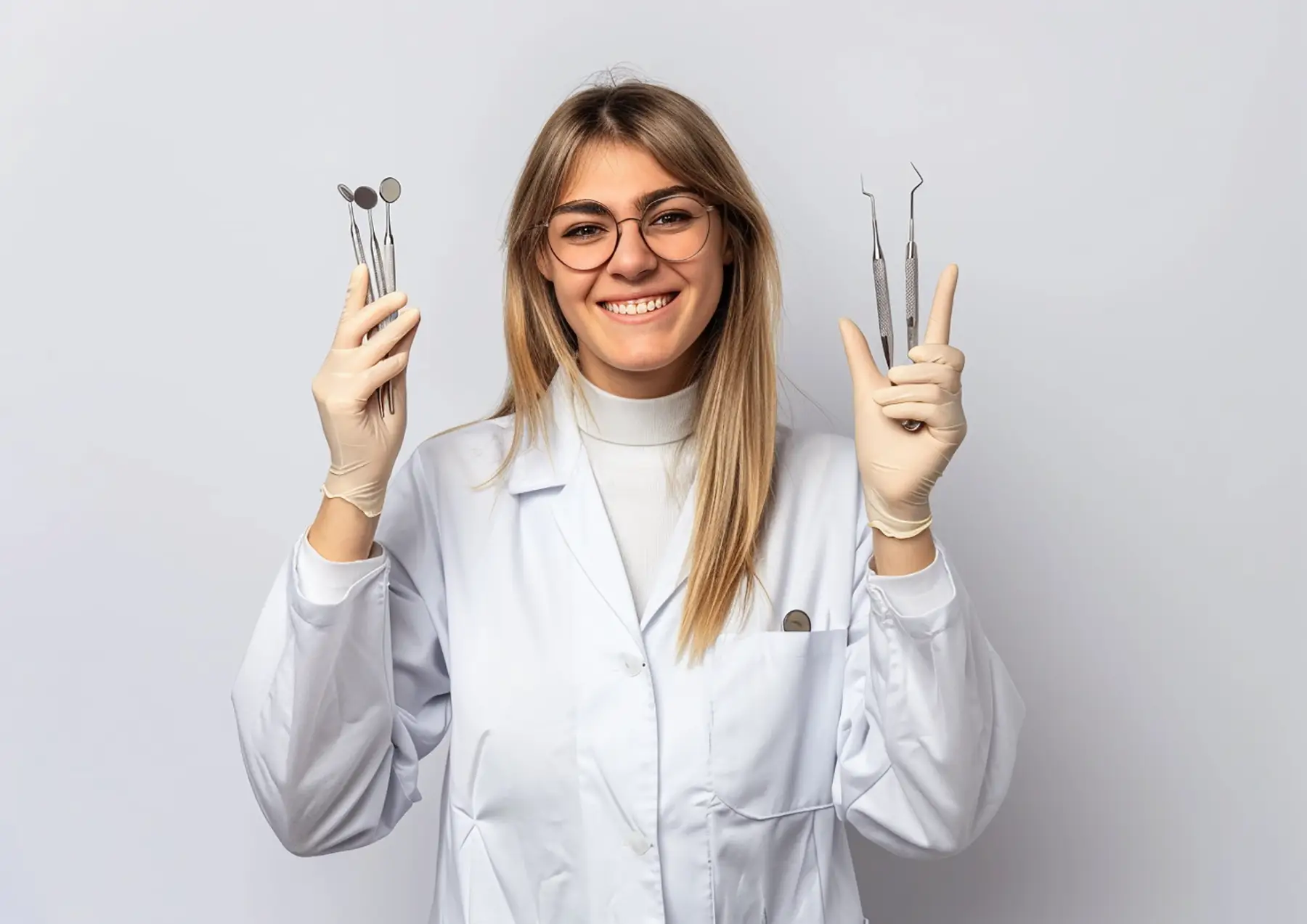 Smiling female dentist wearing glasses and white coat holds dental tools in both gloved hands.