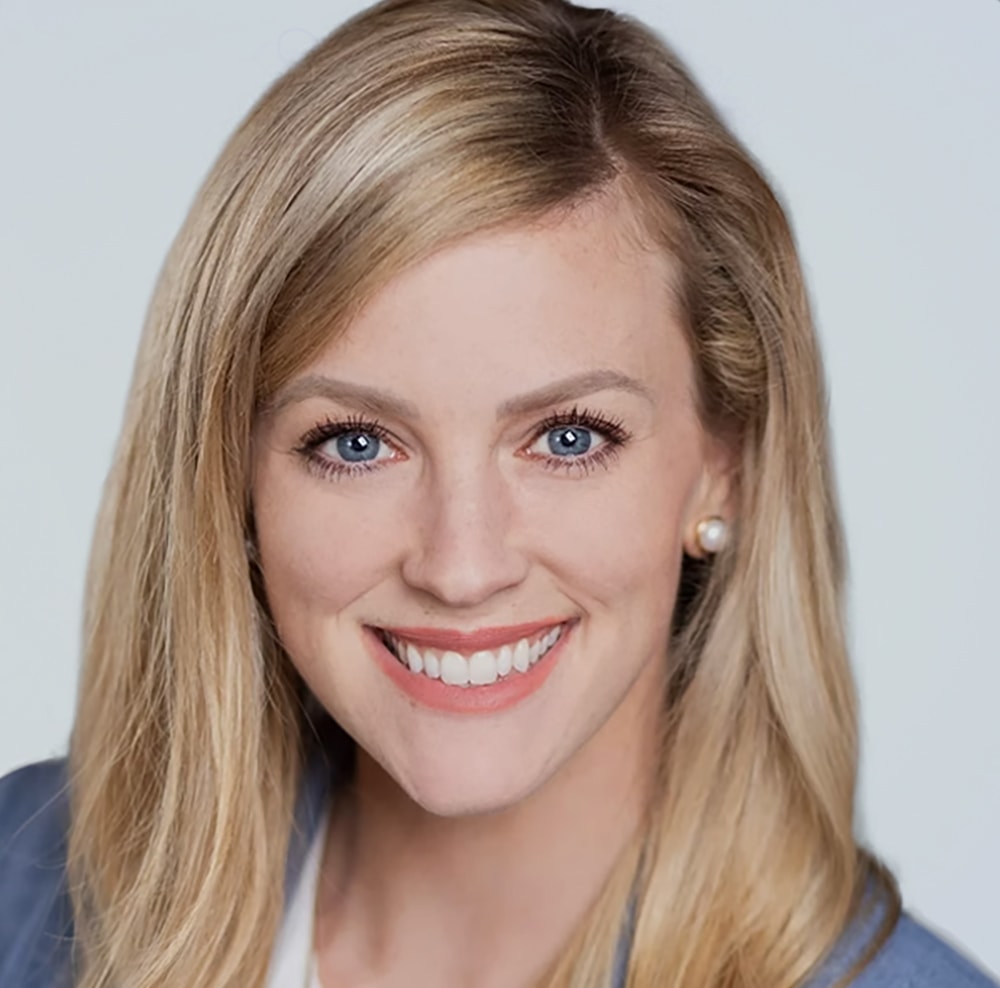 A headshot of a smiling woman with long blonde hair, blue eyes, and wearing a gray blazer. She has pearl earrings and a friendly expression.