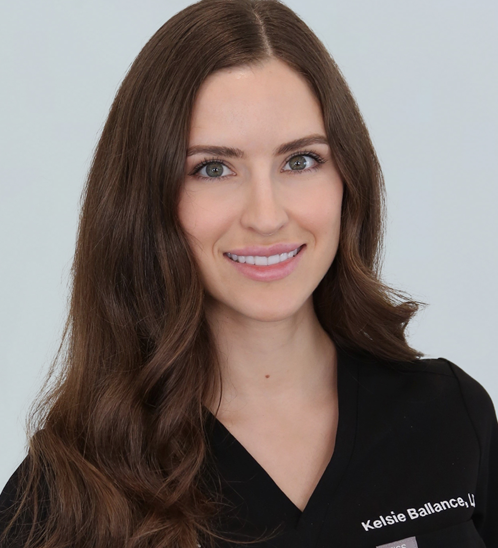 A headshot of a woman with long, wavy brown hair, smiling at the camera while wearing a black medical scrubs top featuring the text 'Kelsie Ballance, LLL' in white.