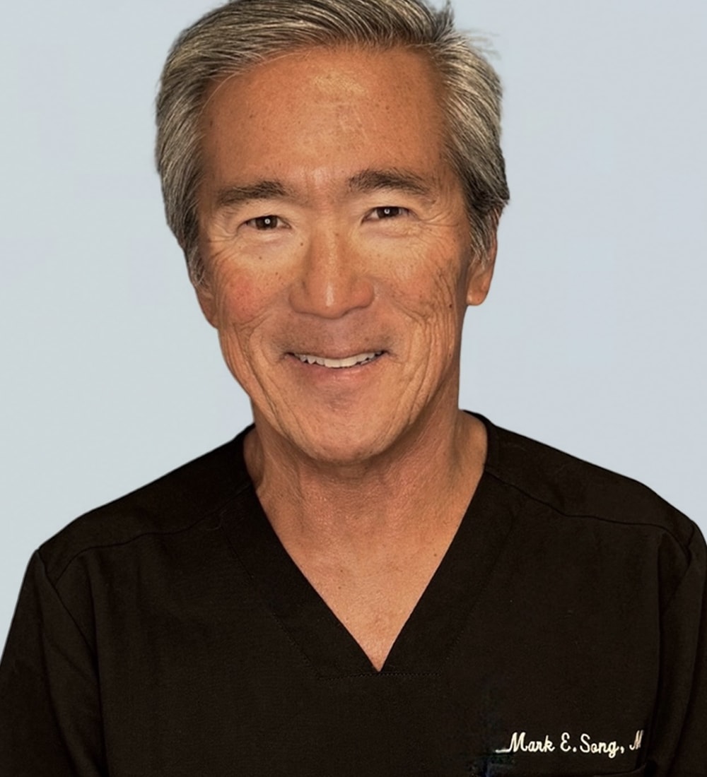 A smiling middle-aged man wearing a black medical scrub top with a sewn name tag that reads 'Mark E. Sang, M.D.'
