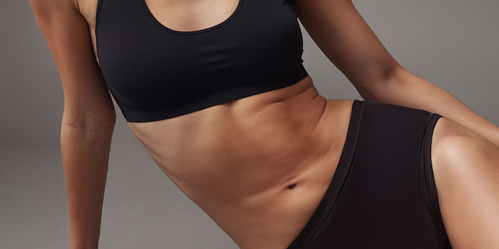 A woman wearing a black sports bra and matching black athletic shorts is exercising with hand weights. Her toned arms and abs are visible, suggesting a focus on strength training and fitness.