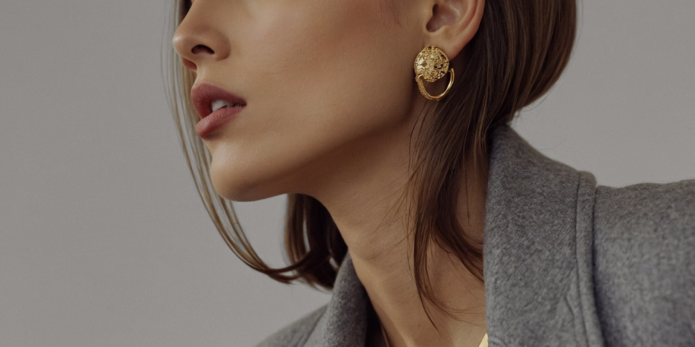 A close-up of a woman’s profile featuring a gold circular earring with a textured design and long, straight hair falling over her shoulder.