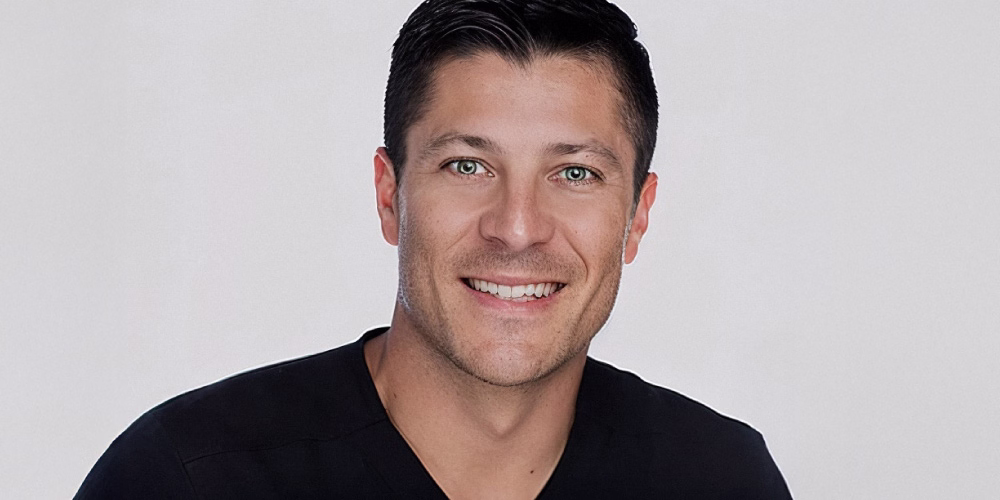 A smiling man with short dark hair and light green eyes, wearing a black shirt. He has a friendly expression and is facing the camera.