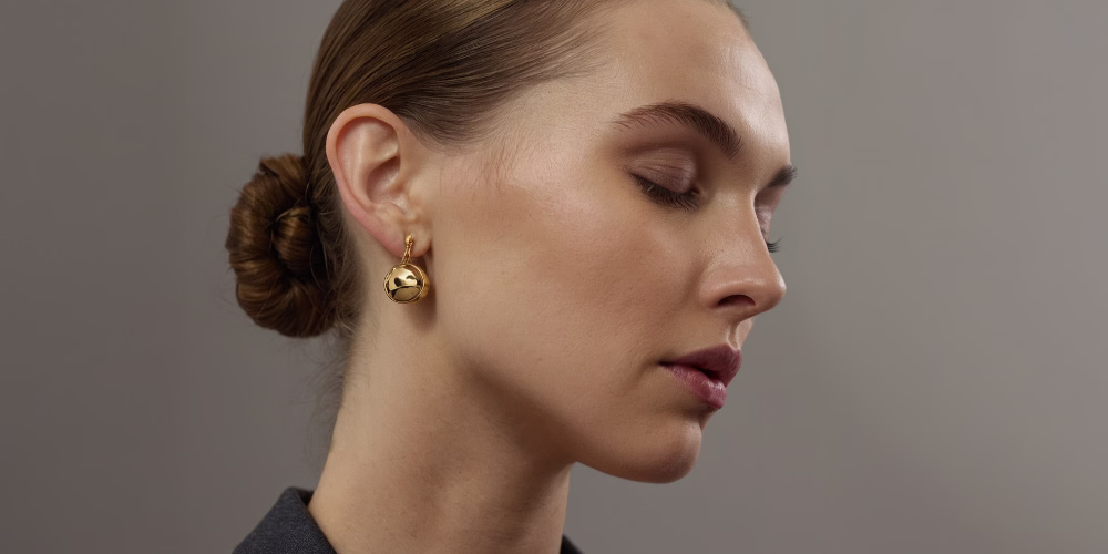 A profile view of a woman with a sleek bun hairstyle, wearing gold hoop earrings and a subtle makeup look.