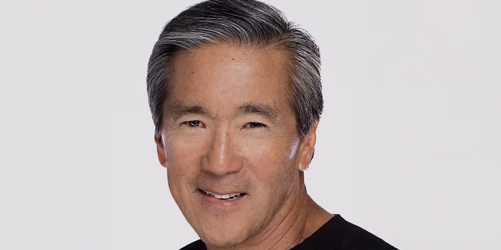 A smiling man with gray hair, wearing a black shirt, looking towards the camera.
